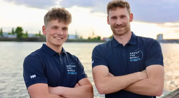 Melwin Fink und sein Teamkollege posieren lächelnd am Wasser. Sie tragen marineblaue Poloshirts mit dem Aufdruck "Around The World - Next Generation".