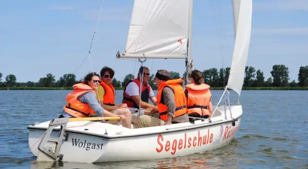 Fünf Personen in orangefarbenen Schwimmwesten genießen das Jolle segeln auf einem sonnigen See.