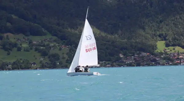 Segel reffen auf einem Boot mit drei Personen. Im Hintergrund bewaldete Hügel mit Häusern.