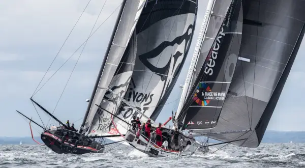Zwei IMOCA 60 Rennyachten kämpfen auf dem Meer, Segel voller Sponsorenlogos und Botschaften für den Klimaschutz.