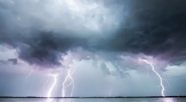 Ein heftiges Gewitter erhellt den Himmel über dem Meer. Blitze zucken, während ein einsames Segelboot auf dem Wasser treibt.