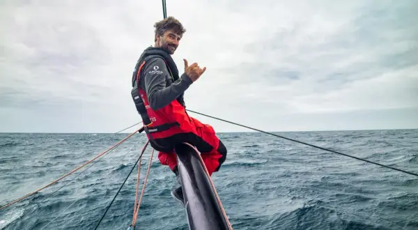 Boris Herrmann winkt lässig vom Bug seines Bootes, umgeben von rauer See und Segeltrossen.