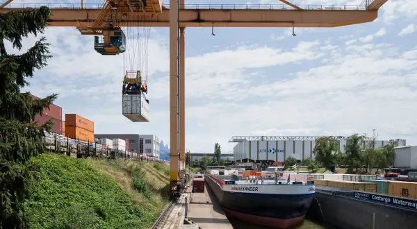 Ein Kran hebt einen Container im Hafen der Schweiz. Ein Frachtschiff liegt am Kai, umgeben von weiteren Containern und Lagerhallen.