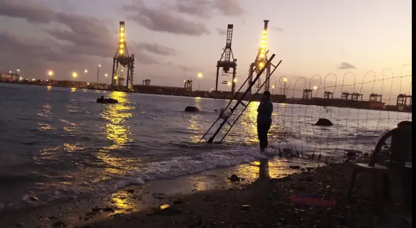 Abendstimmung am Hafen von Port Sudan. Kräne leuchten im Wasser, eine Person steht am Ufer.