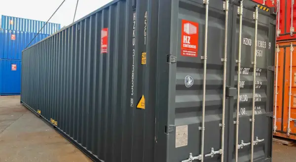 Ein grauer soc container wird von einer Kette angehoben. Daneben stehen blaue und rote Container.