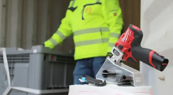 Werkzeug für Container Lashing: Ein Mann in Warnweste lädt Kisten, während ein Akkuschrauber und ein Cutter bereitliegen.
