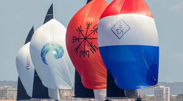 Segelboote der j class rasen über das Meer. Bunte Spinnaker mit Logos und Symbolen, im Hintergrund eine Küstenstadt.