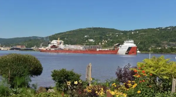 Der Frachter Paul R. Tregurtha fährt auf einem ruhigen Fluss, umgeben von grünen Hügeln und blühenden Blumen.