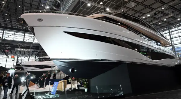 Eine glänzende weiße Luxus-**Boot**-Yacht mit dunklen Fenstern, ausgestellt auf einer Messe. Im Hintergrund eine weitere Yacht und Besucher.