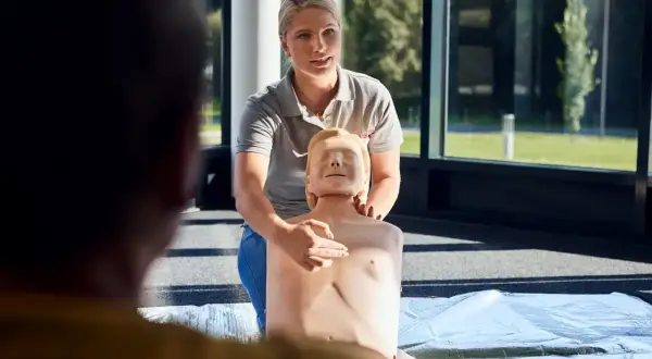 Frau zeigt an einer Übungspuppe die Herzmassage. Sie erklärt die korrekte Position für den Williamson Turn.