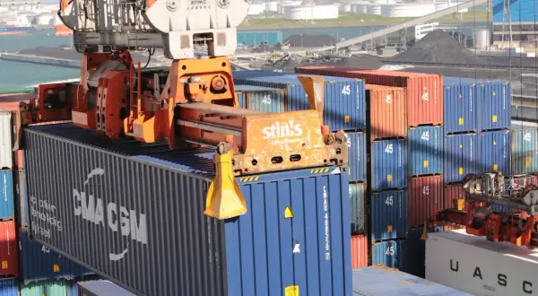 Ein Kran hebt einen blauen CMA CGM-Container. Viele weitere Container stapeln sich im Hafen, kein Container ist unberührt.