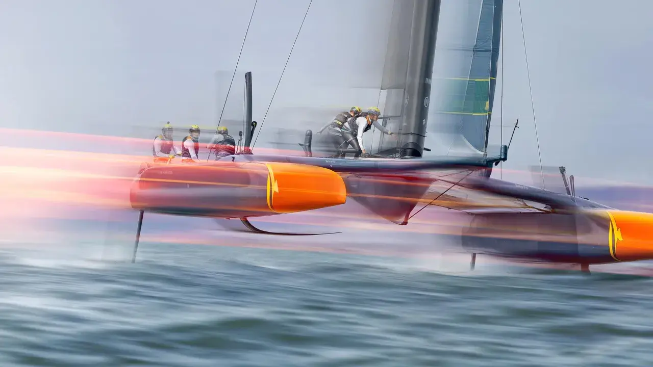 Schnelles Katamaran-Segelboot mit Besatzung, das über das Wasser gleitet. Die orangefarbenen Rümpfe und die Segel sind durch die Bewegung verschwommen, was die Geschwindigkeit des Sail GP-Rennens zeigt.