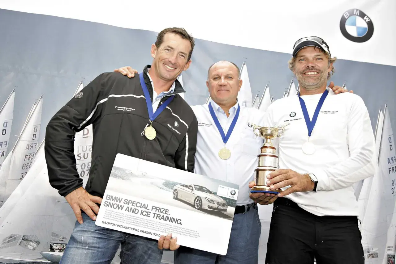 Markus Wieser mit zwei Segelkollegen und einem Pokal. Sie halten eine Broschüre für BMW Snow and Ice Training.