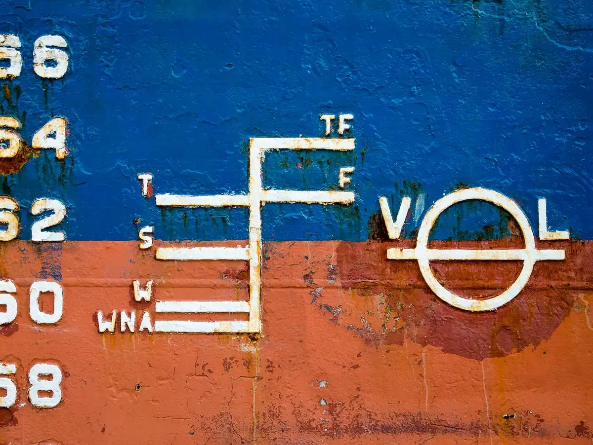 Schiffsbord mit Markierungen, die die Tiefgangslinien (plimsoll line) anzeigen. Zahlen und Buchstaben wie 56, 54, 52, 50, 68, T, S, W, WNA, TF, F, V, O, L sind sichtbar.