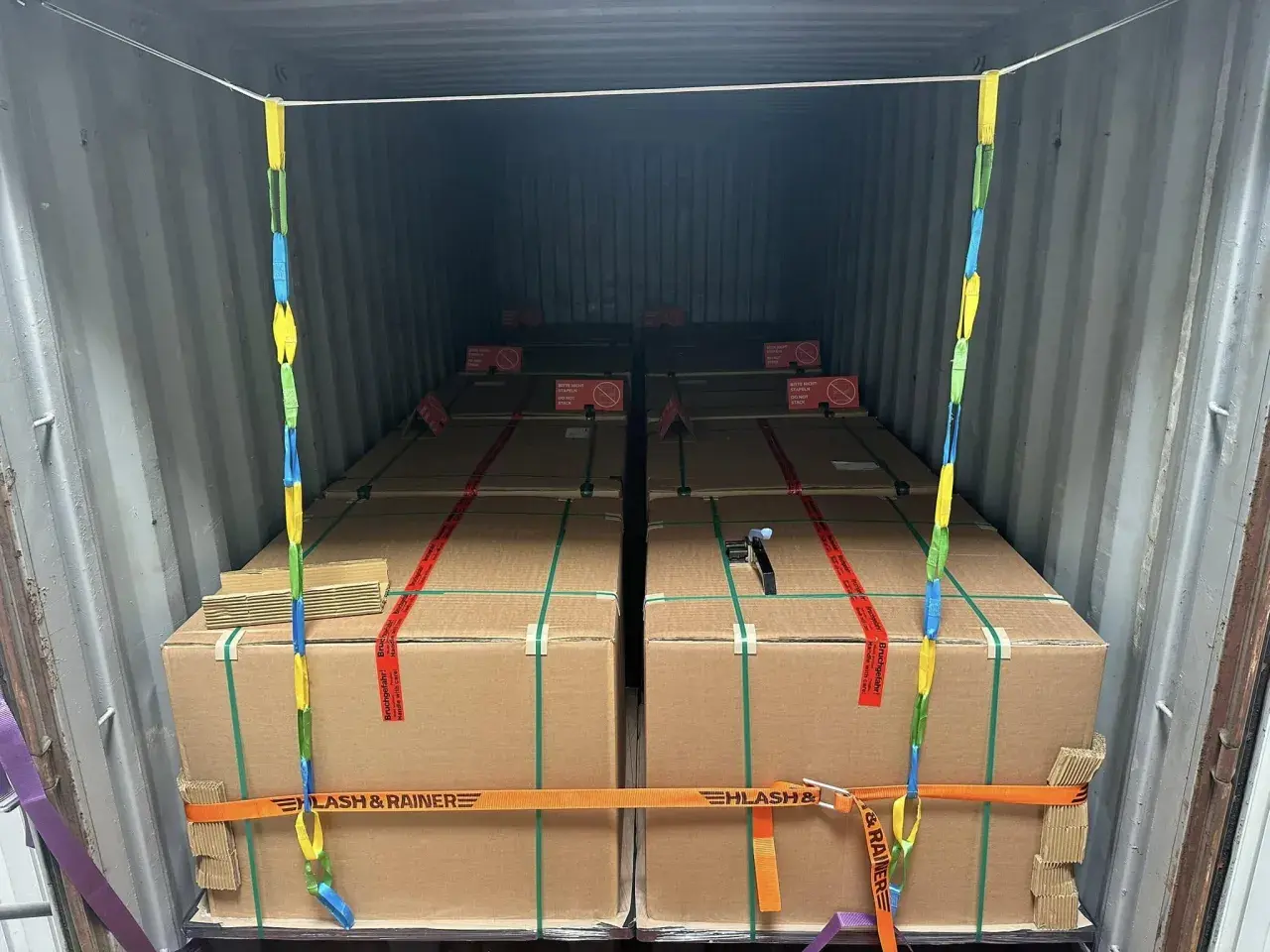 Ladungssicherung in einem Frachtcontainer: Mehrere große Kartons, gesichert mit Gurten und Ketten. Verschiedene Arten von Containern werden hier für den Transport genutzt.