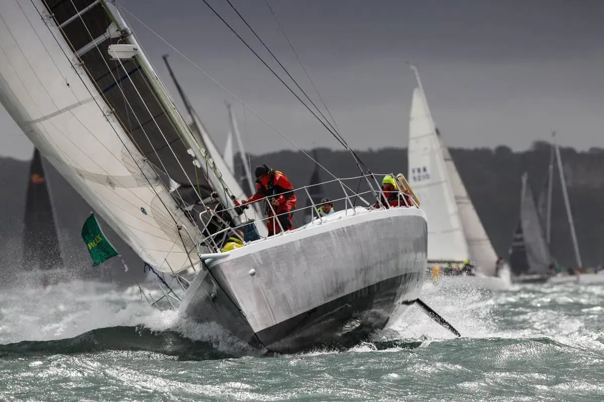 Segelboote kämpfen sich durch raue Wellen beim Fastnet Race. Ein Segler in Rot hält sich fest, während die Gischt aufspritzt.