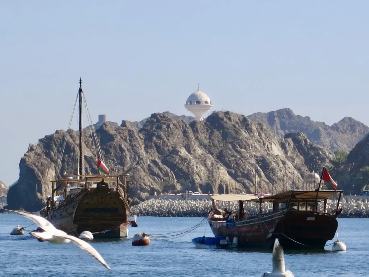 Traditionelle Dhaus-Boote im Oman Port, mit felsigen Bergen und einem weißen Kuppelgebäude im Hintergrund.