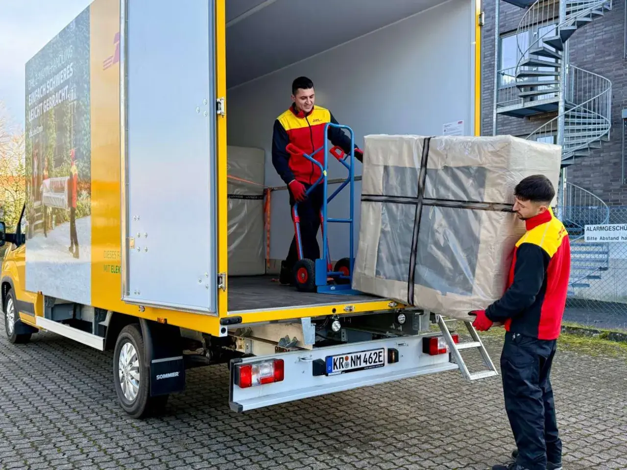 Zwei Männer laden ein großes Paket aus einem LKW. Ein Mann schiebt es mit einem Hubwagen, der andere trägt es. Maersk e-delivery macht es möglich.
