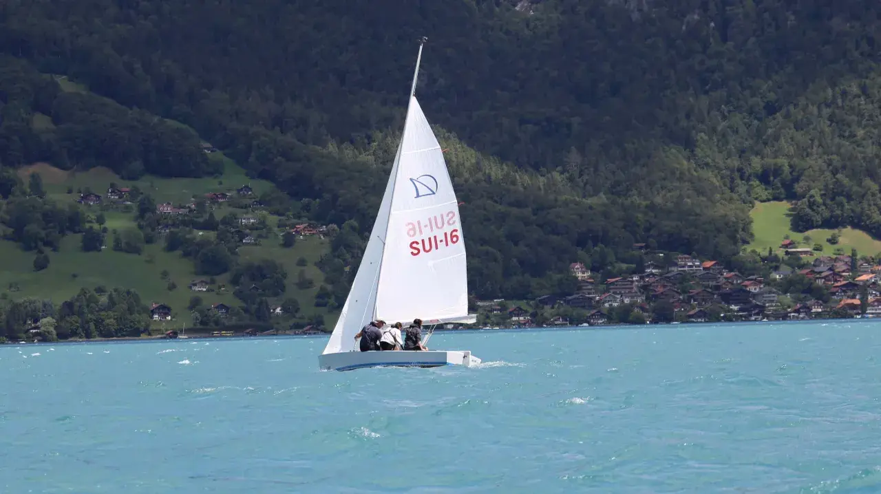 Segel reffen auf einem Boot mit drei Personen. Im Hintergrund bewaldete Hügel mit Häusern.