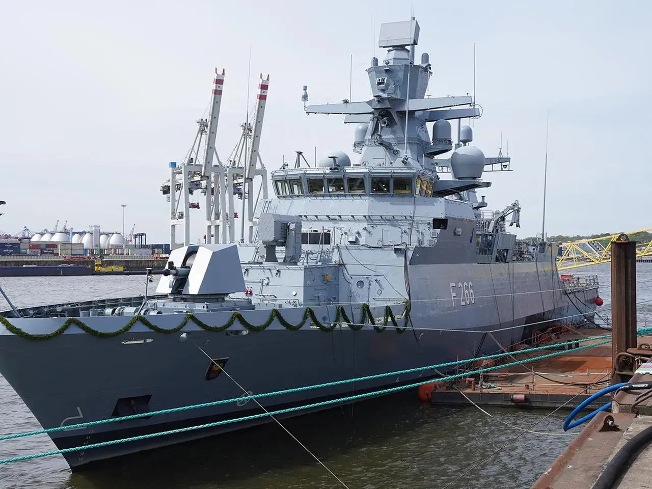 Graue corvette F266 im Hafen, mit Kranen und Containern im Hintergrund.