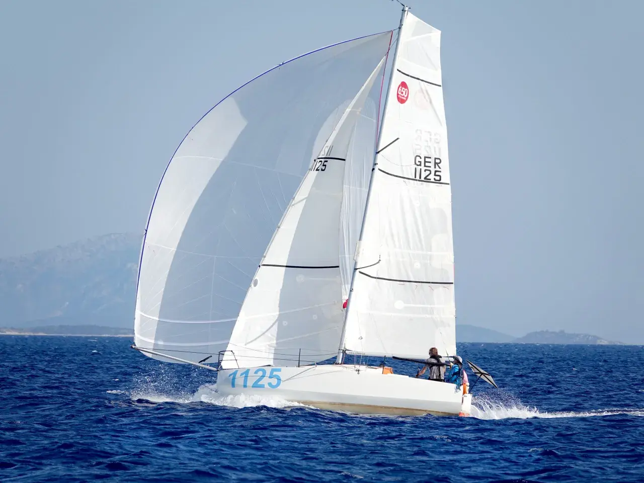 Segelboot mit zwei Personen an Bord, das auf dem Meer fährt. Die Segel sind weiß und mit der Nummer 1125 beschriftet. Dies ist ein Bild von einem Mini Transat Rennen.