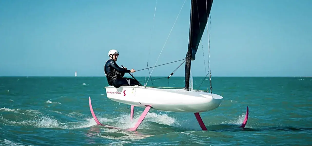Ein Mann in Neoprenanzug und Helm steuert ein Boot mit pinken Flügeln, das über das Wasser gleitet. Das ist modernes Foil Segeln.