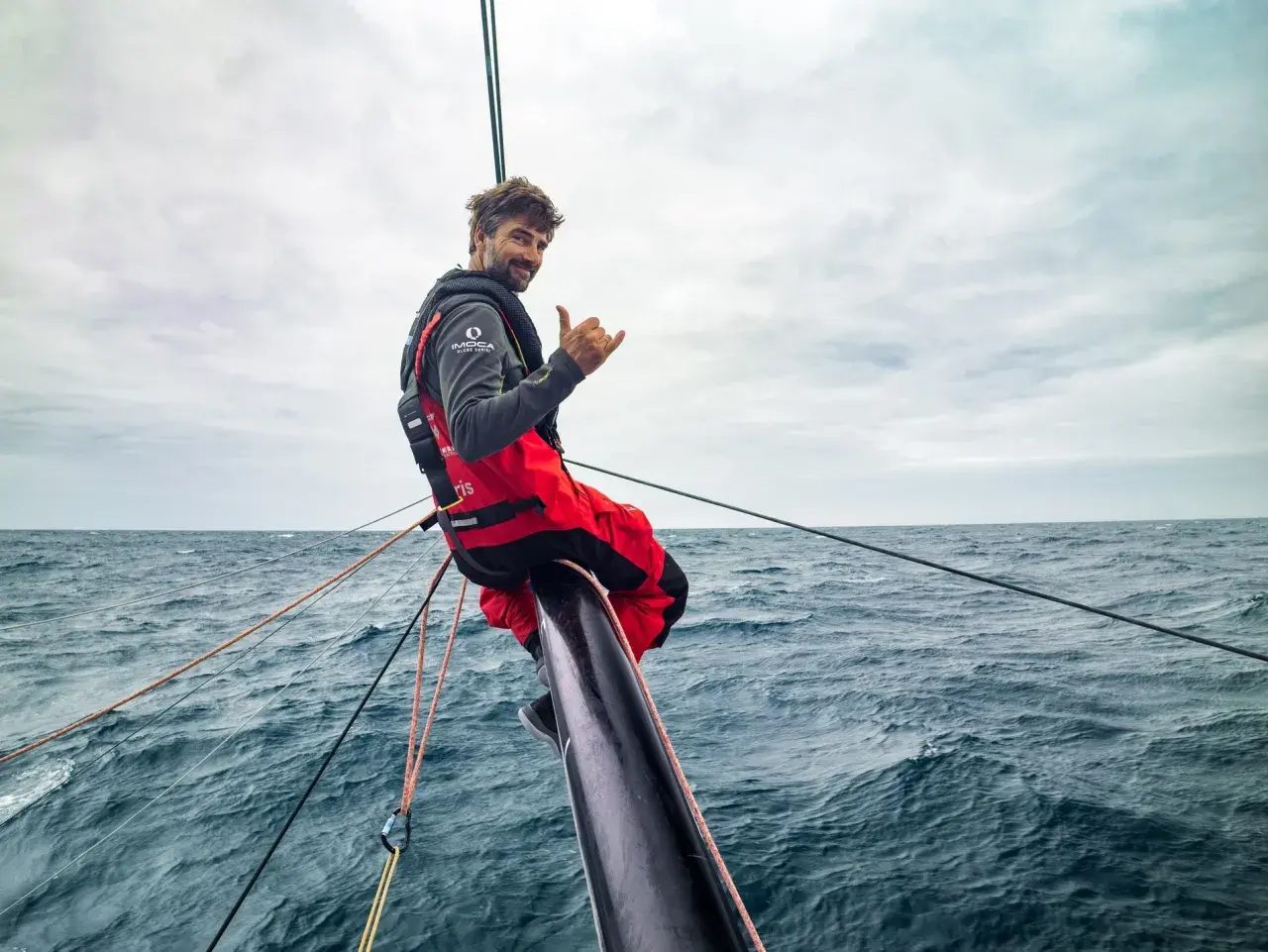 Boris Herrmann winkt lässig vom Bug seines Bootes, umgeben von rauer See und Segeltrossen.