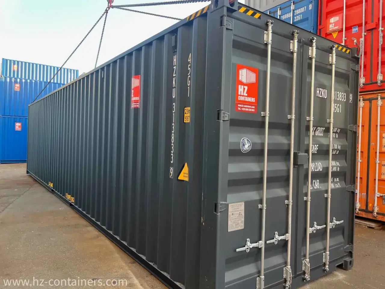Ein grauer soc container wird von einer Kette angehoben. Daneben stehen blaue und rote Container.