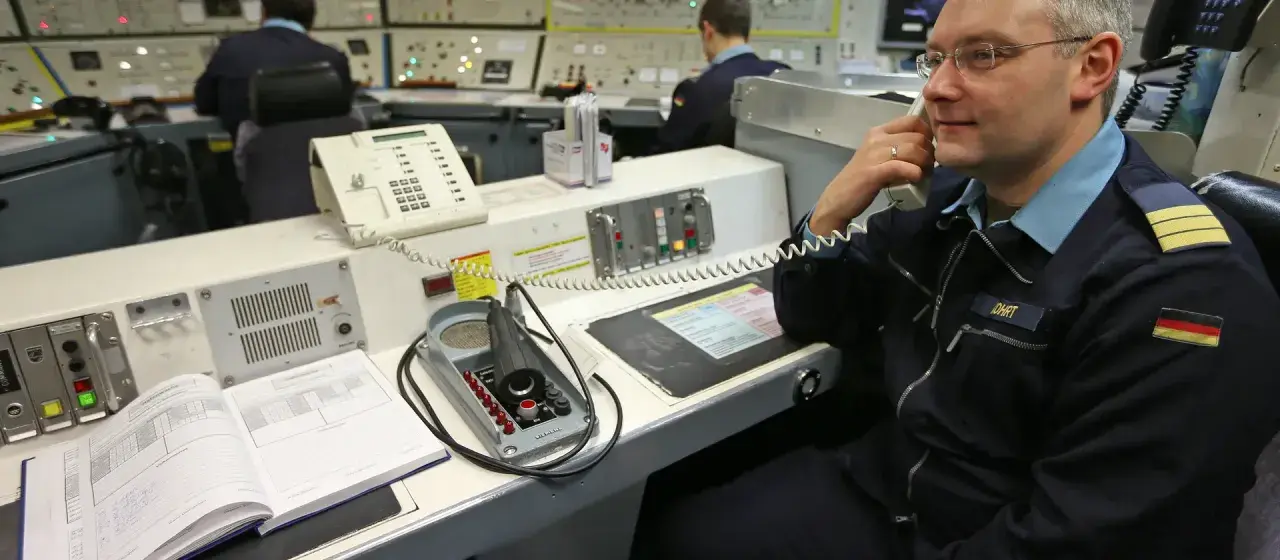 Mann im Marine-Outfit telefoniert in einem Kontrollraum. Ein offenes Buch und ein Mikrofon liegen auf dem Tisch. Marine-Ingenieurwesen im Einsatz.