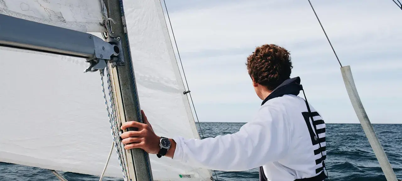 Ein junger Mann mit lockigem Haar blickt aufs Meer, während er die Hand am Mast hält. Er ist bereit, die Segel zu setzen und in See zu stechen.