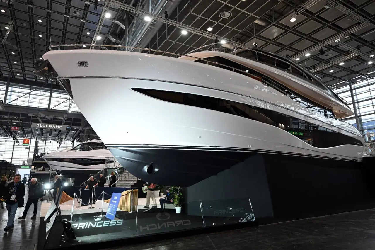 Eine glänzende weiße Luxus-**Boot**-Yacht mit dunklen Fenstern, ausgestellt auf einer Messe. Im Hintergrund eine weitere Yacht und Besucher.