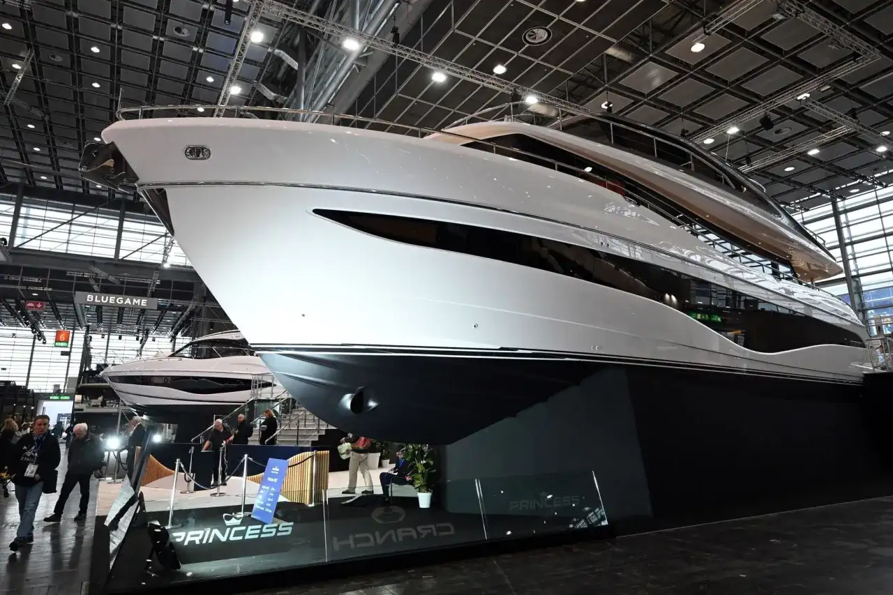 Eine glänzende weiße Luxus-**Boot**-Yacht mit dunklen Fenstern, ausgestellt auf einer Messe. Im Hintergrund eine weitere Yacht und Besucher.