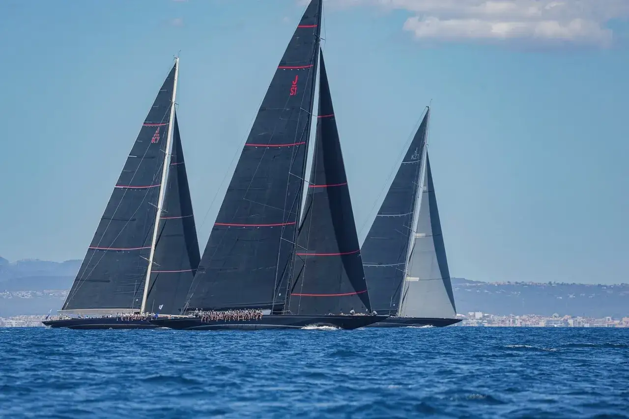 Drei J-Klasse Yachten segeln auf dem Meer. Die schwarzen Segel mit roten Streifen sind imposant.