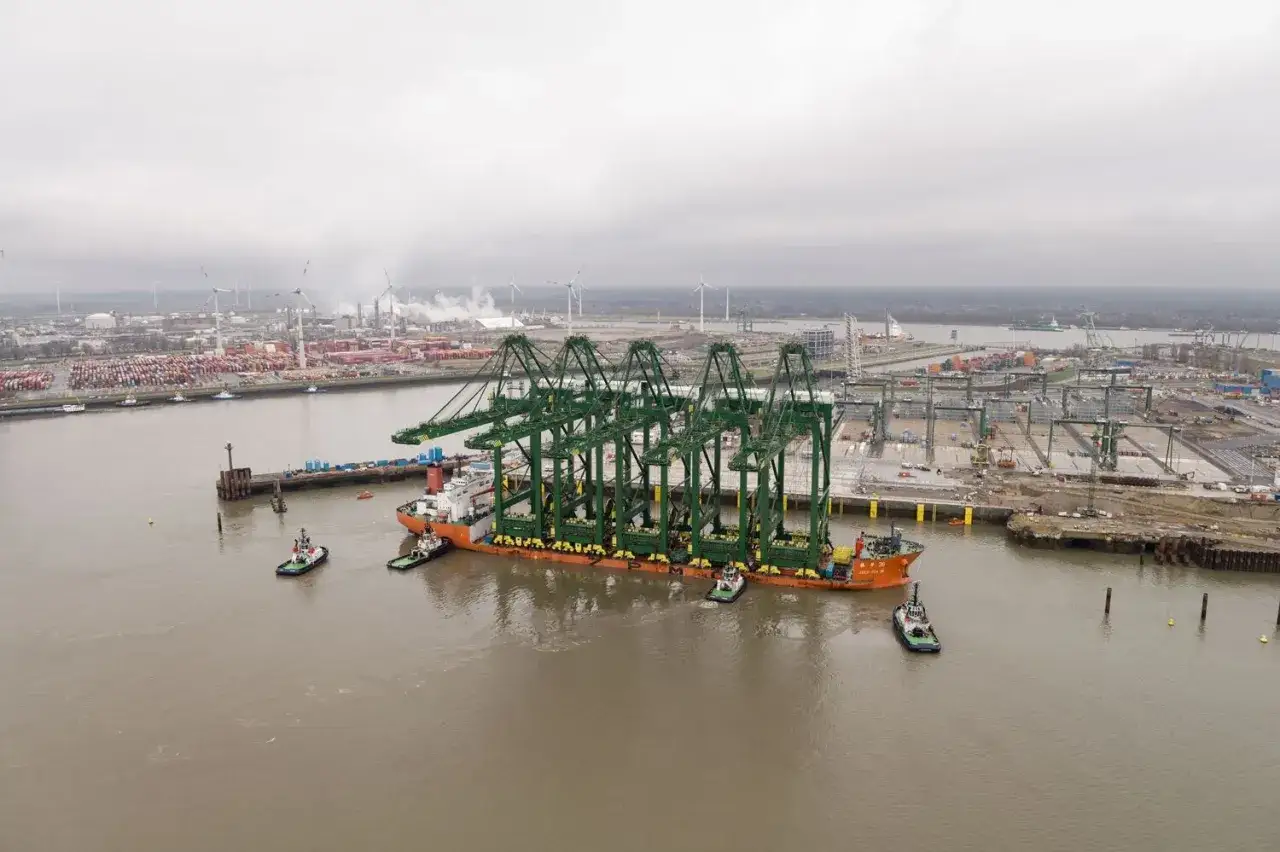 Schwere Kräne werden im Antwerpen Port auf einem Schiff transportiert, umgeben von Schleppern und der weitläufigen Hafenanlage.