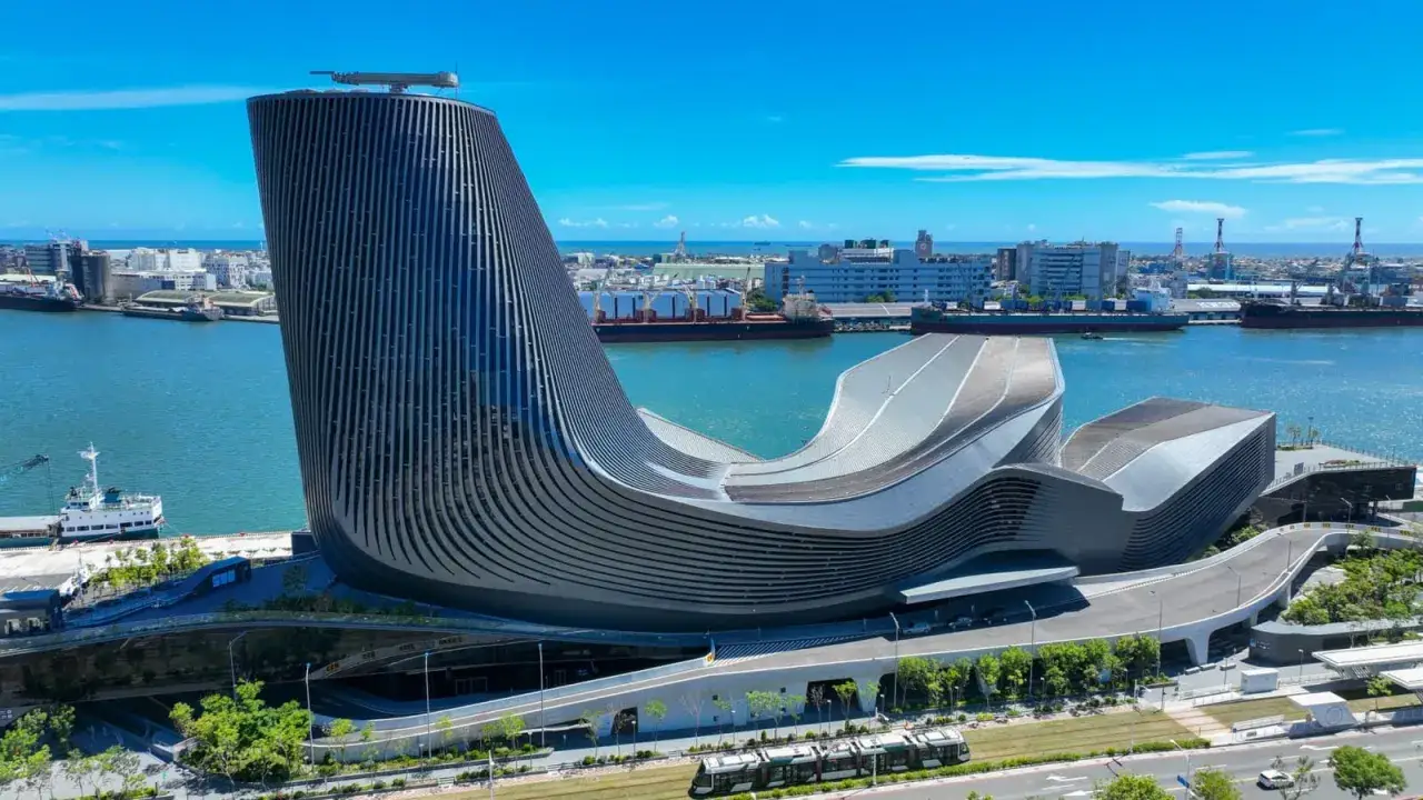 Moderne Architektur am Kaohsiung Port: Ein geschwungenes Geb&auml;ude mit vertikalen Lamellen ragt neben Frachtschiffen und dem blauen Meer auf.