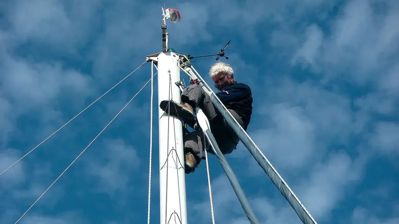 Mann klettert am Mast eines Segelboots in den Himmel.