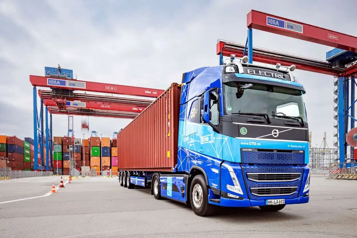Blauer Elektro-LKW mit rotem Container im Hafen, bereit zur Abholung durch den Empf&auml;nger.
