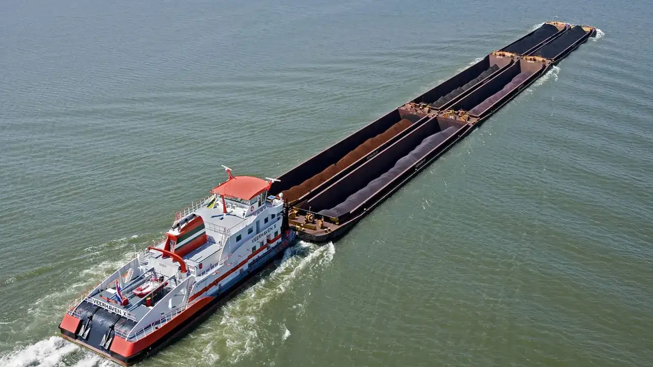 Ein Schlepper zieht ein langes barge ship mit Kohle beladen &uuml;ber das Wasser.