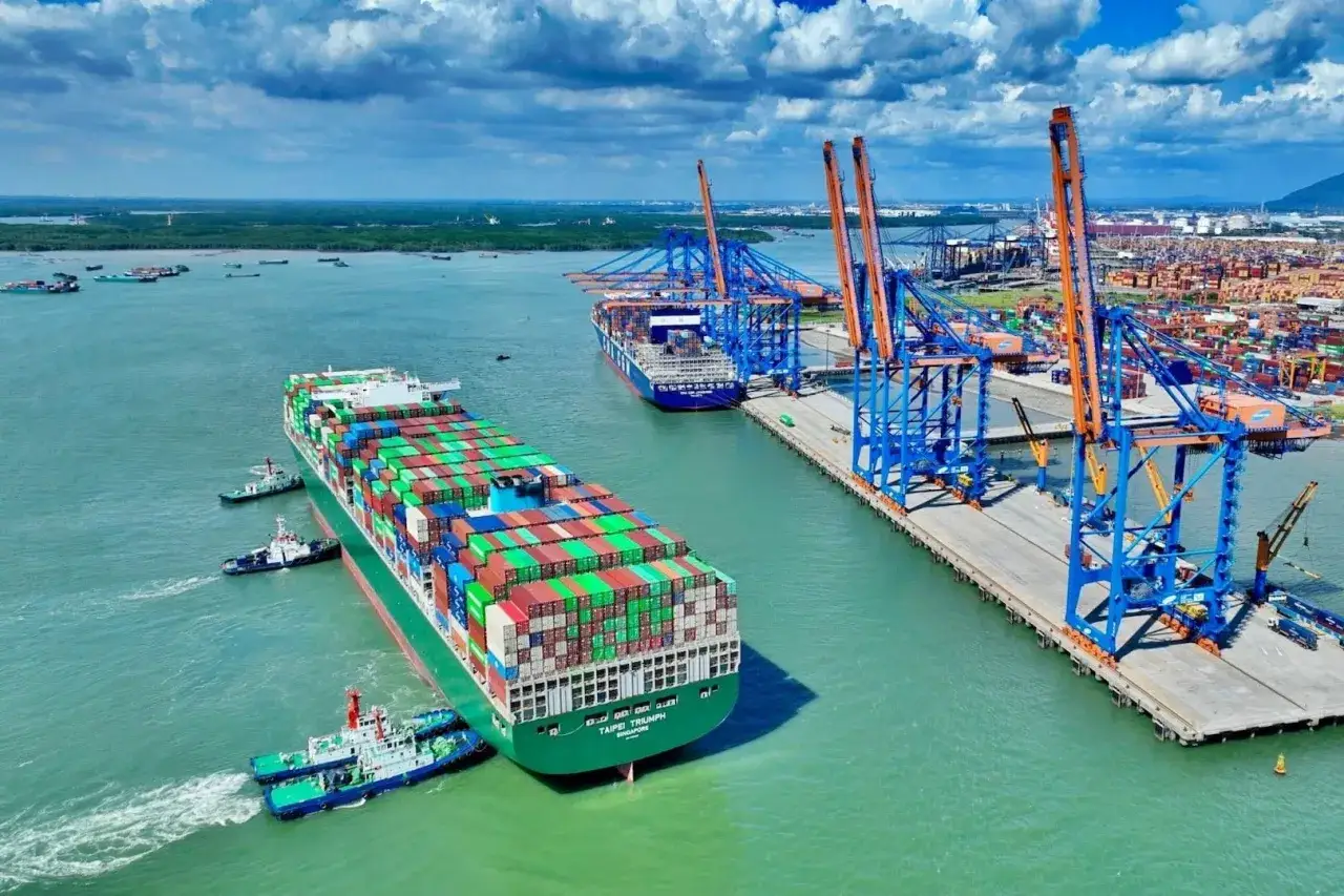 Großes Containerschiff wird von Schleppern in einem geschäftigen Cat Lai Port manövriert, umgeben von Kränen und anderen Schiffen.