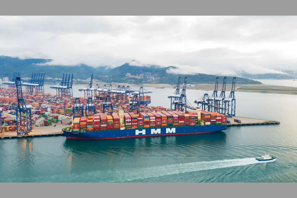 Ein riesiges Containerschiff der HMM-Reederei liegt im Hafen, beladen mit unzähligen Frachtcontainern. Kräne ragen in den Himmel, bereit für die nächste Ladung.