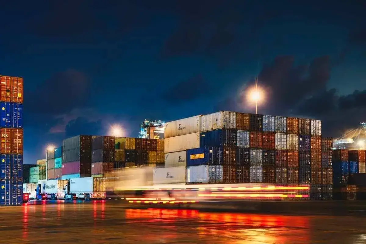 Nachts im Oman Port: Ein LKW f&auml;hrt durch Reihen von Frachtcontainern, deren Lichter sich auf dem nassen Boden spiegeln.
