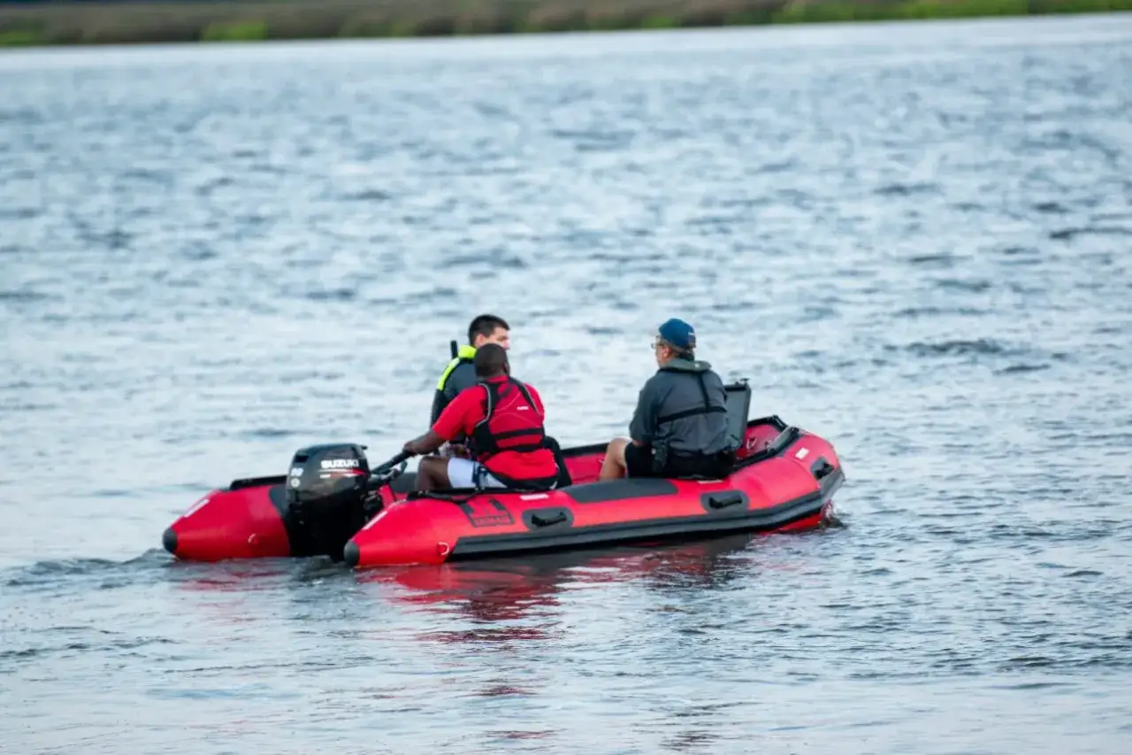 Rotes Schlauchboot mit drei M&auml;nnern, die auf dem Wasser fahren. Einer steuert, die anderen schauen. Ein Mann tr&auml;gt eine Rettungsweste. Das Boot scheint Wasser aufzunehmen, als ob es sinken w&uuml;rde.