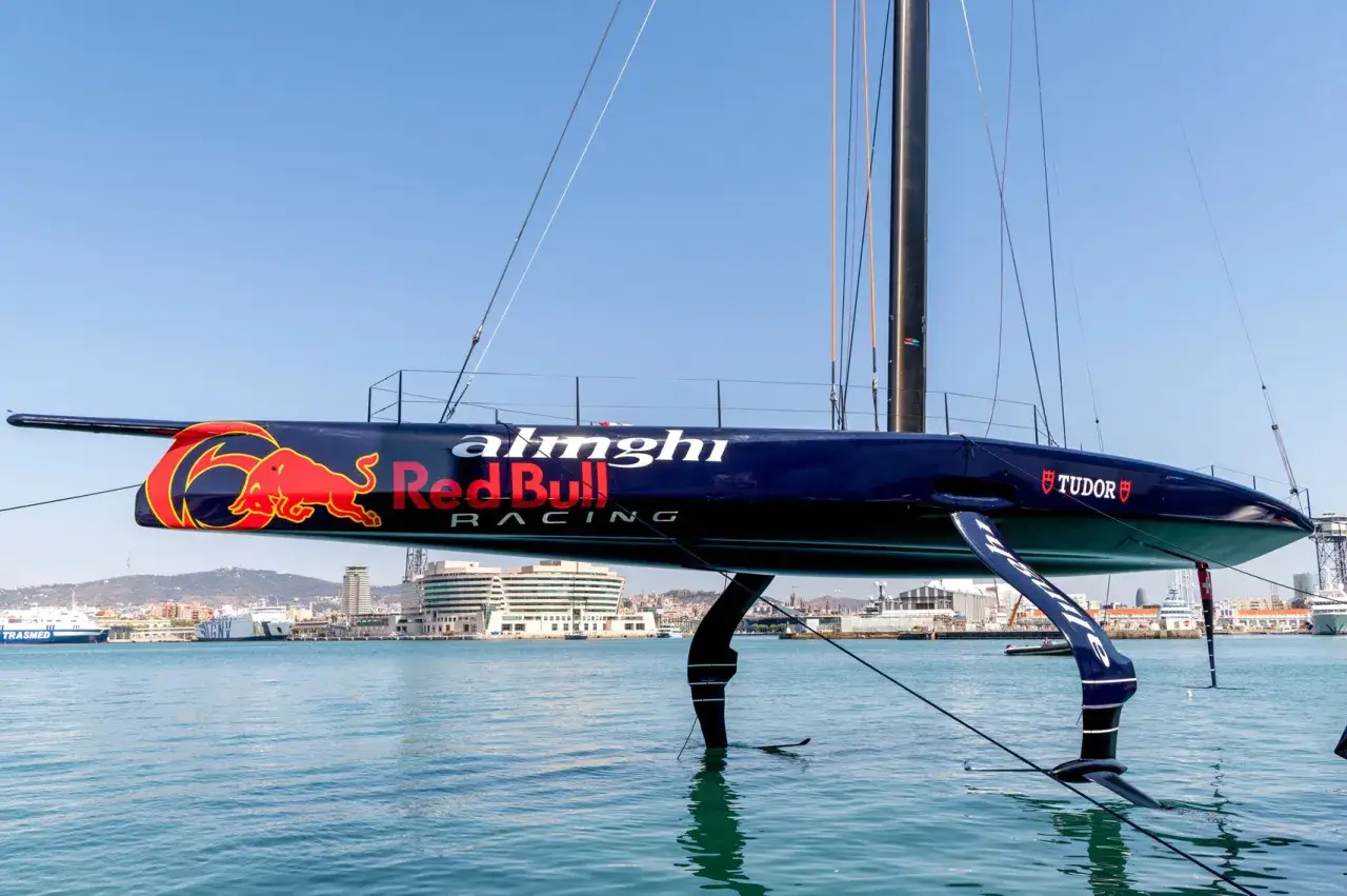 Der Alinghi Red Bull Racing Katamaran hebt sich mit seinen Foils aus dem Wasser. Ein roter Bulle ziert das dunkle Schiff.