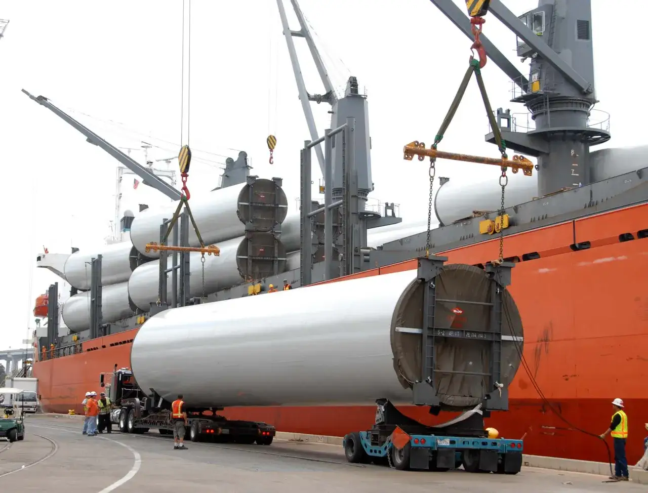 Große Windturbinenteile werden per break bulk-Verfahren von einem Schiff entladen. Kräne heben die langen Zylinder auf Tieflader.