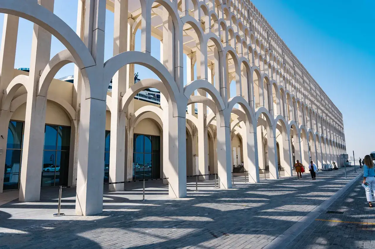 Ein langer Arkadengang mit vielen B&ouml;gen am doha port. Menschen spazieren auf dem gepflasterten Weg entlang der modernen Architektur.
