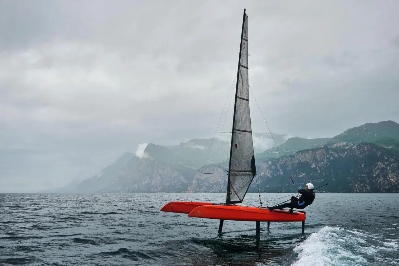 Ein Segler auf einem orangefarbenen Katamaran mit Foils gleitet über das Wasser. Die Berge im Hintergrund sind in Nebel gehüllt.