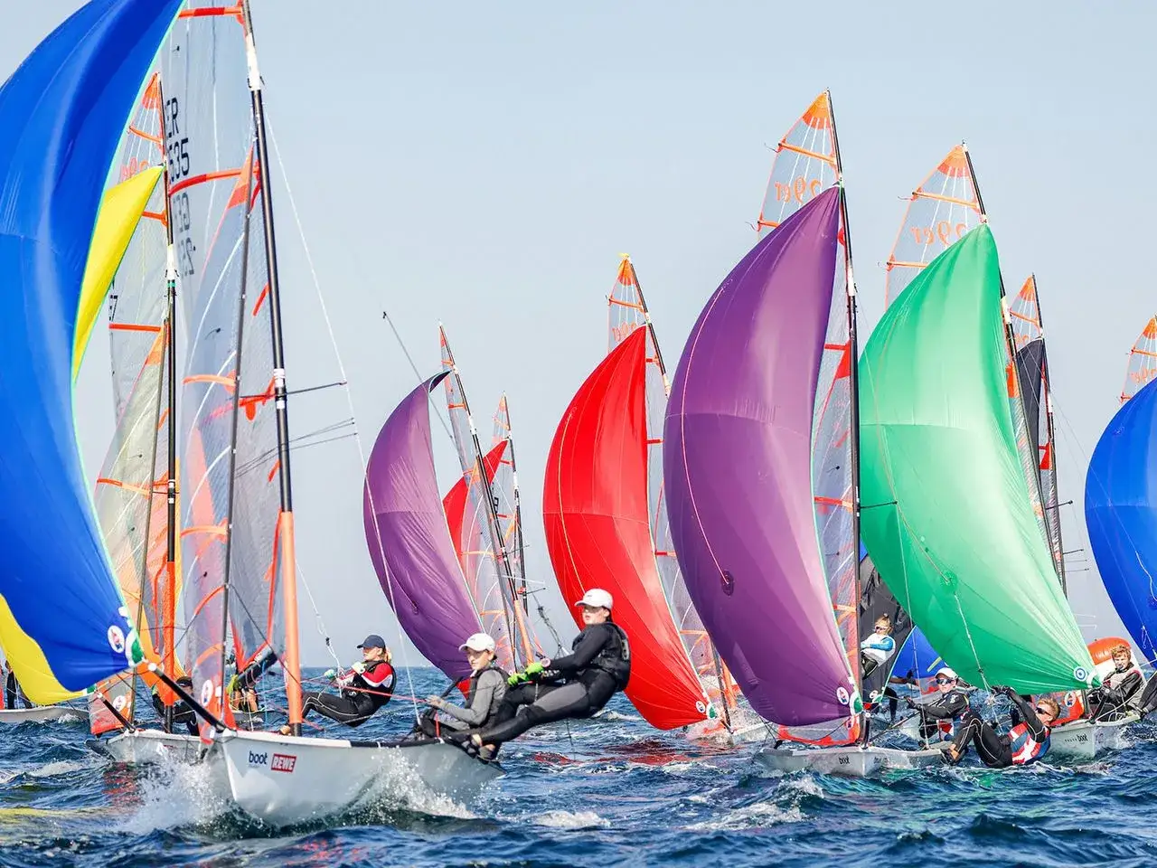 Bunte Segel dominieren das Bild beim Kieler Woche Segeln. Mehrere Boote mit Crews kämpfen auf dem Wasser um die Wette.
