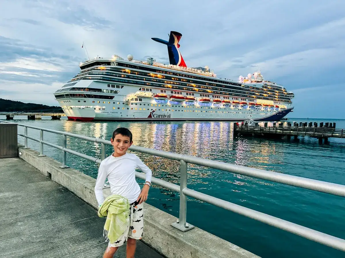 Junge steht vor der Carnival Freedom. Das Kreuzfahrtschiff leuchtet im Abendlicht, ein Feuerwerk der Lichter spiegelt sich im Wasser.