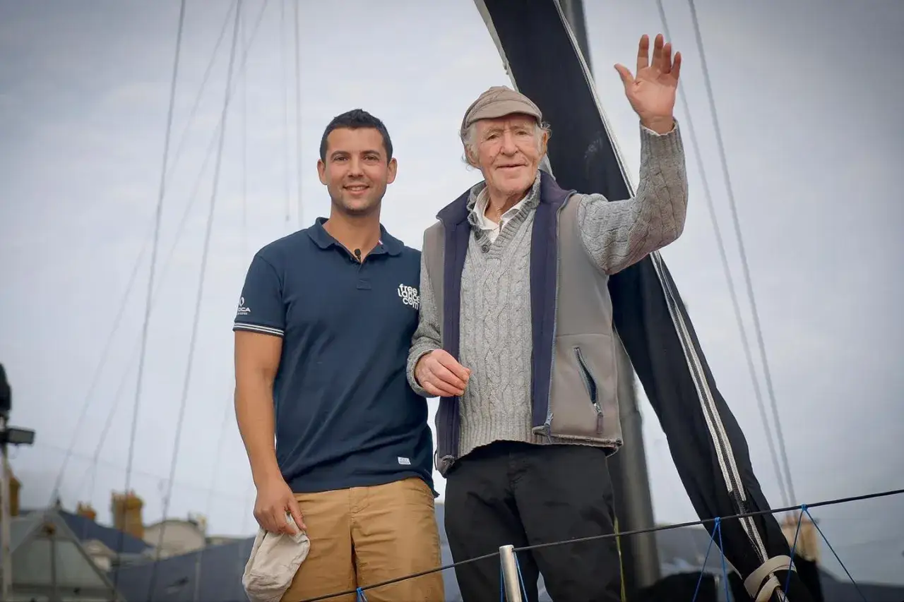 Zwei Männer auf einem Boot. Der ältere, mit Hut und winkend, ist Guirec Soudée.