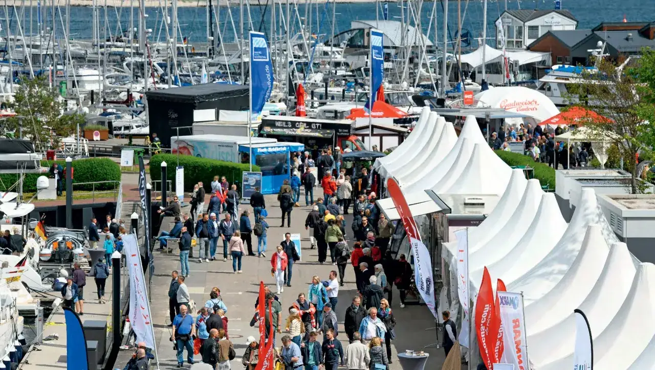 Besucher bummeln über die ancora boat show, vorbei an weißen Zelten und vielen Segelbooten im Hafen.